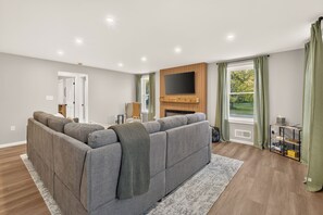 Een moderne woonkamer met een hoekbank, een flatscreen televisie bevestigd aan een houten paneel en een open haard.