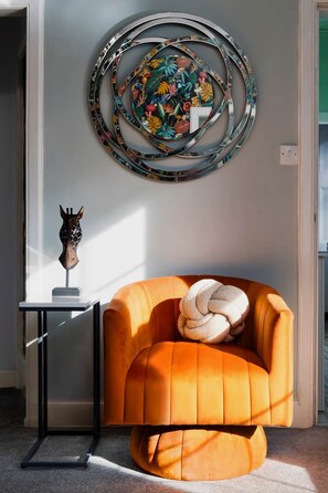 Ein modernes Wohnzimmer mit einem leuchtend orangefarbenen Sessel, einem runden Wandbild und einer kleinen Skulptur auf einem Beistelltisch.