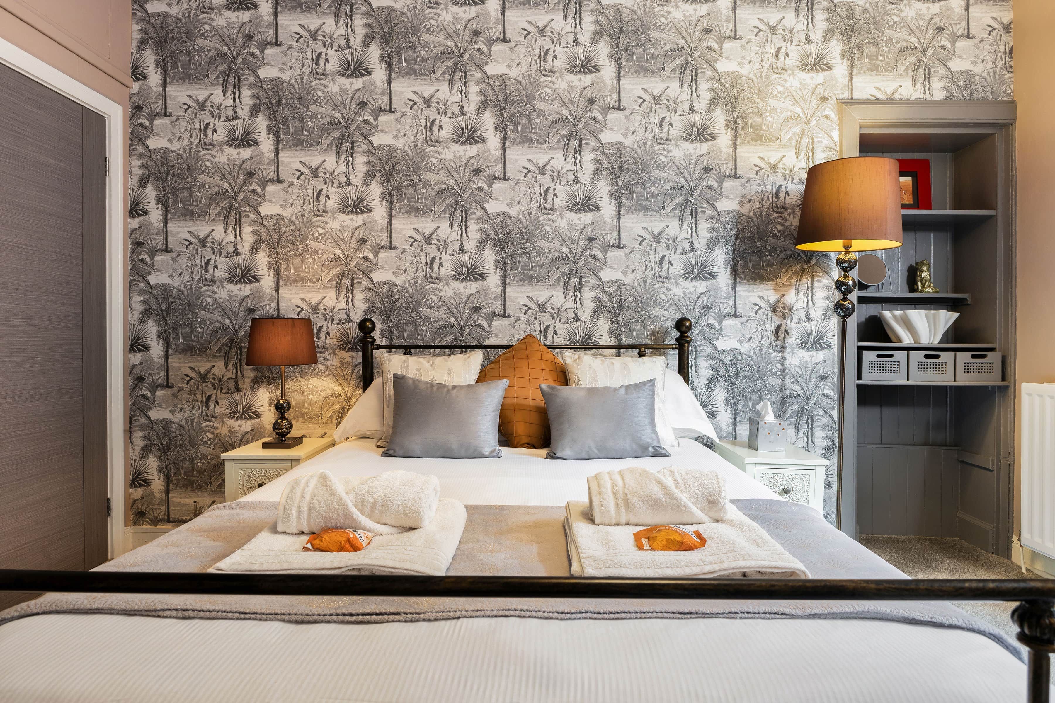 A hotel room with a bed, two bedside lamps, a nightstand, a shelf with decorative items, and a wall with a tropical wallpaper pattern.