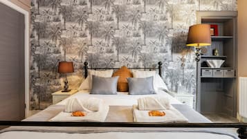 A hotel room with a bed, two bedside lamps, a nightstand, a shelf with decorative items, and a wall with a tropical wallpaper pattern.