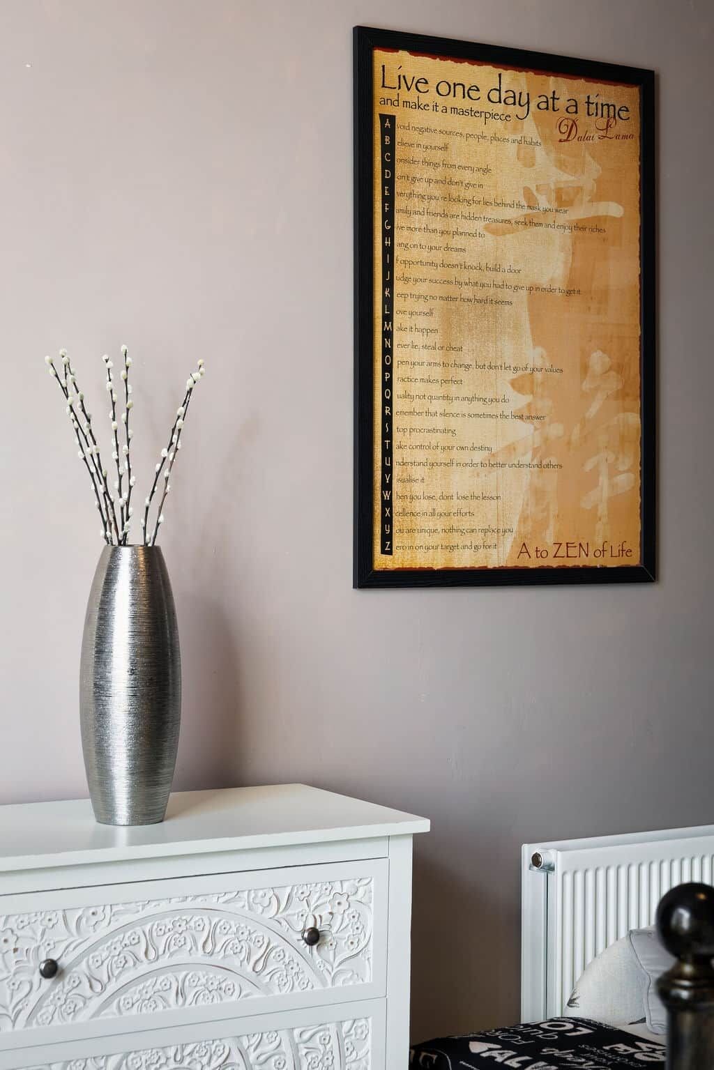 Um quadro com uma frase inspiradora na parede, um vaso decorativo com galhos e uma cômoda branca com desenhos detalhados.