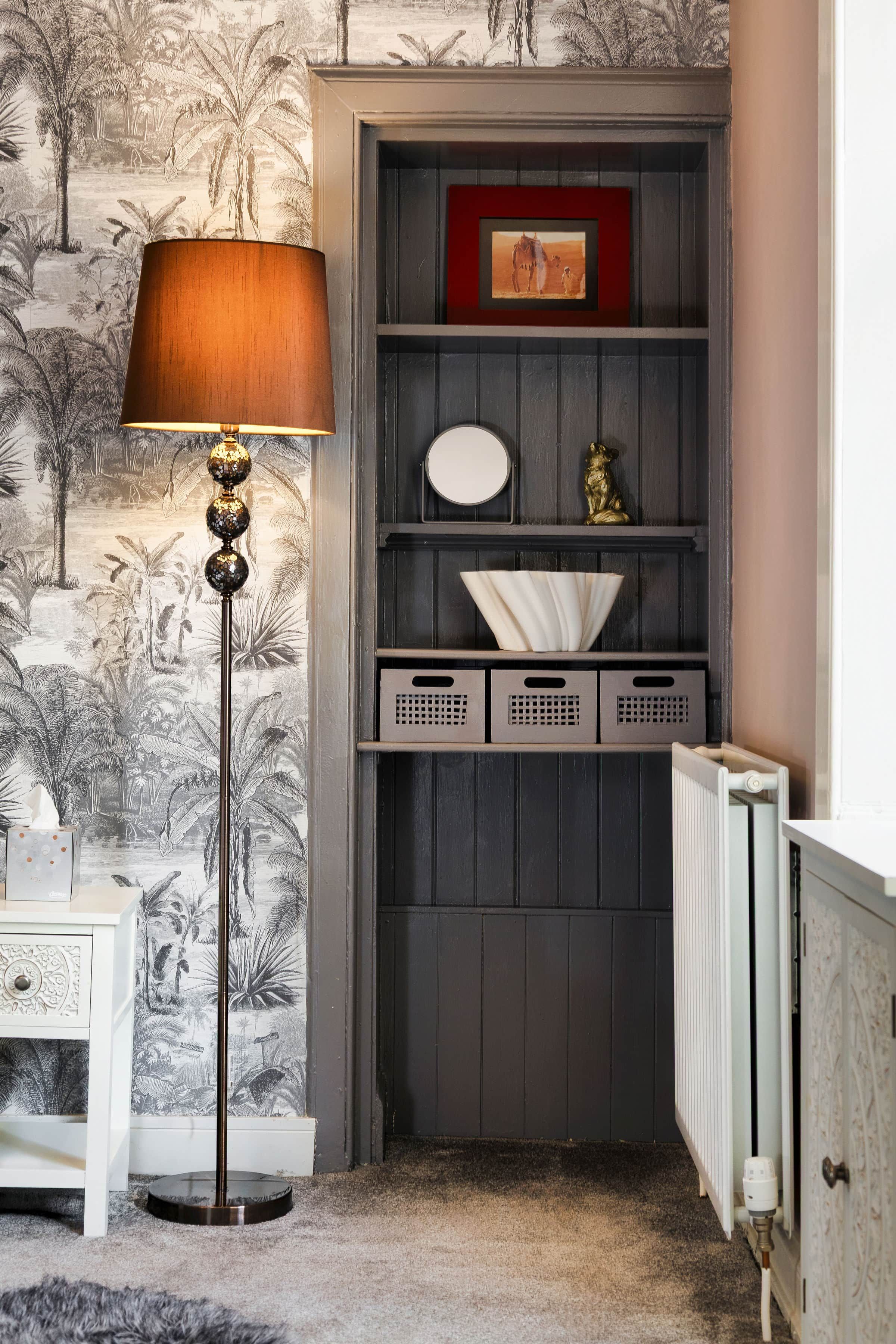 A room with a radiator, a floor lamp, a shelf with decorative items, and a wall with a tropical wallpaper.