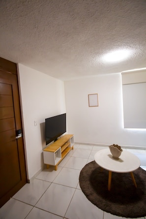 Ein Zimmer mit einem Flachbildfernseher, einem Holz-TV-Schrank, einem runden Tisch mit einer Vase und einer Tür.