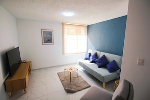 A living room with a sofa, a television, a small wooden table, and a window with blinds. - Dptos. Las Rosas Expo Guadalajara (Guadalajara)