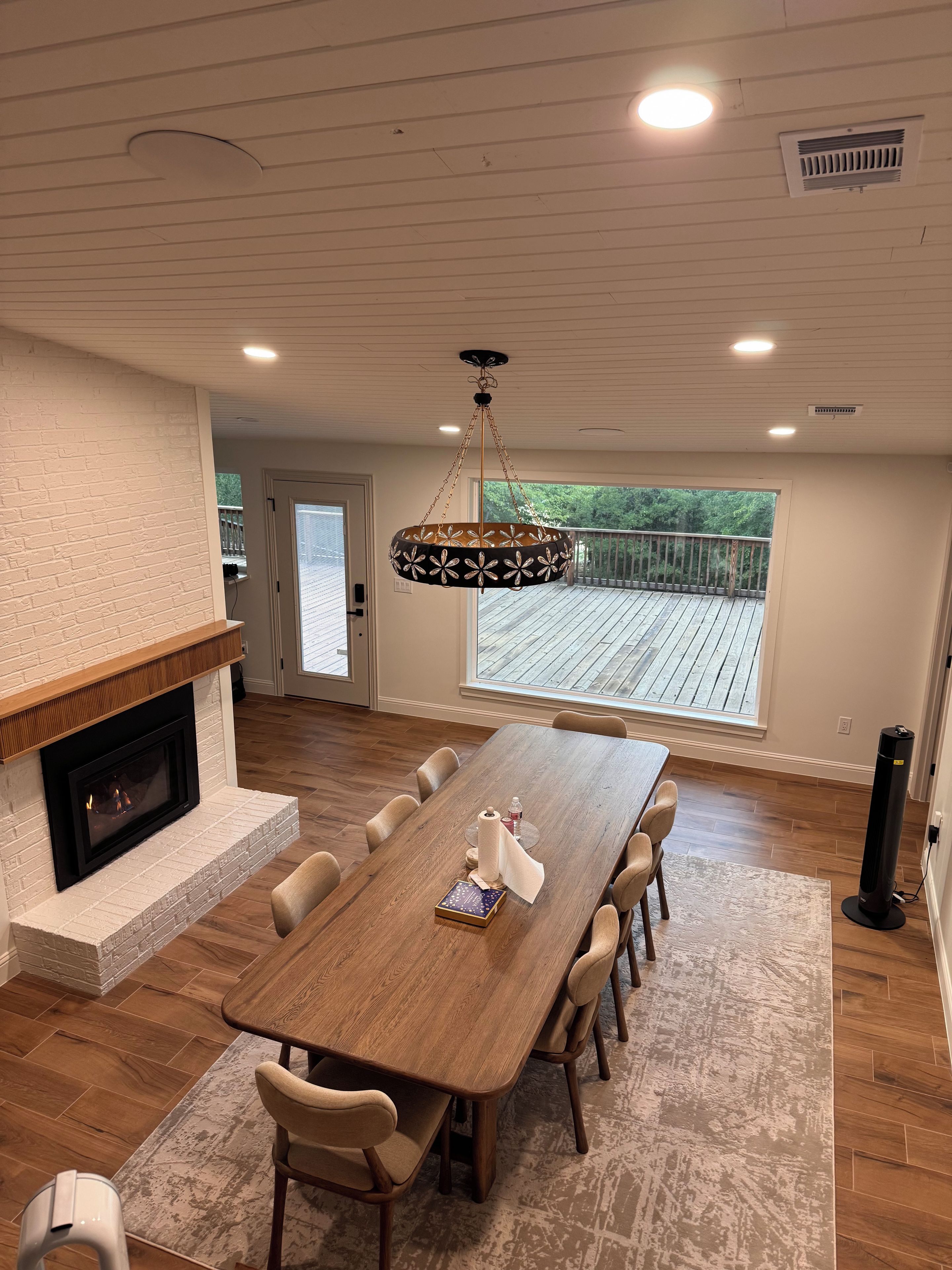 A spacious dining area with a long wooden table, comfortable chairs, a fireplace, and a view of a wooden deck through a large window.