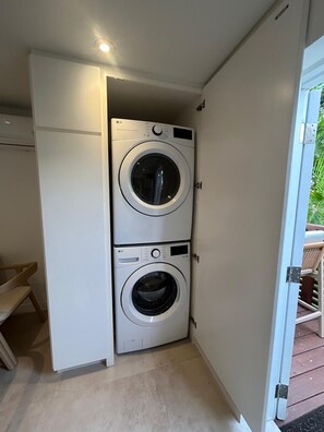 A laundry room with two stacked washing machines, a bench, and a door leading to an outdoor area. - Atlantic Shores - 1 Bedroom Apartment (Christ Church)