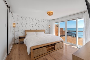 Un dormitorio con una cama grande, cabecero de madera y vistas al mar a través de un ventanal corredero.