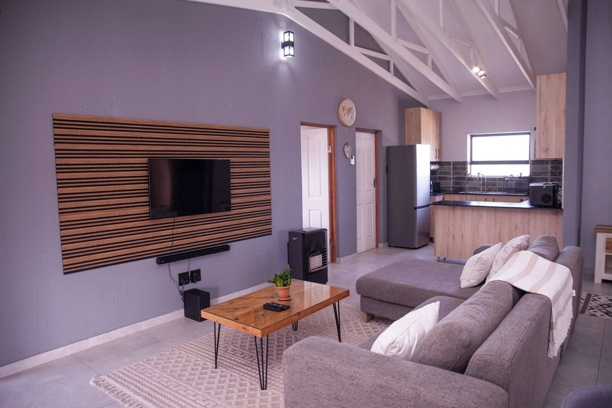 Una sala moderna con un televisor de pantalla plana montado en un panel de madera, un sofá, una mesa de centro y una cocina al fondo.