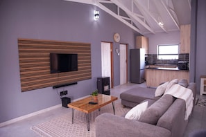 A modern living room with a flat-screen TV mounted on a wooden panel, a sofa, a coffee table, and a kitchen area in the background. - Langa Elihle on Paradise Beach (Paradise Beach)