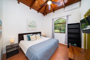 A bedroom with a bed, a nightstand, a wall-mounted TV, a ceiling fan, a window with curtains, and a door. - Pool ~ BBQ ~ Walk to Beach ~ Golf Cart Parking ~ 3 TVs - Villaggio 324 (Potrero)