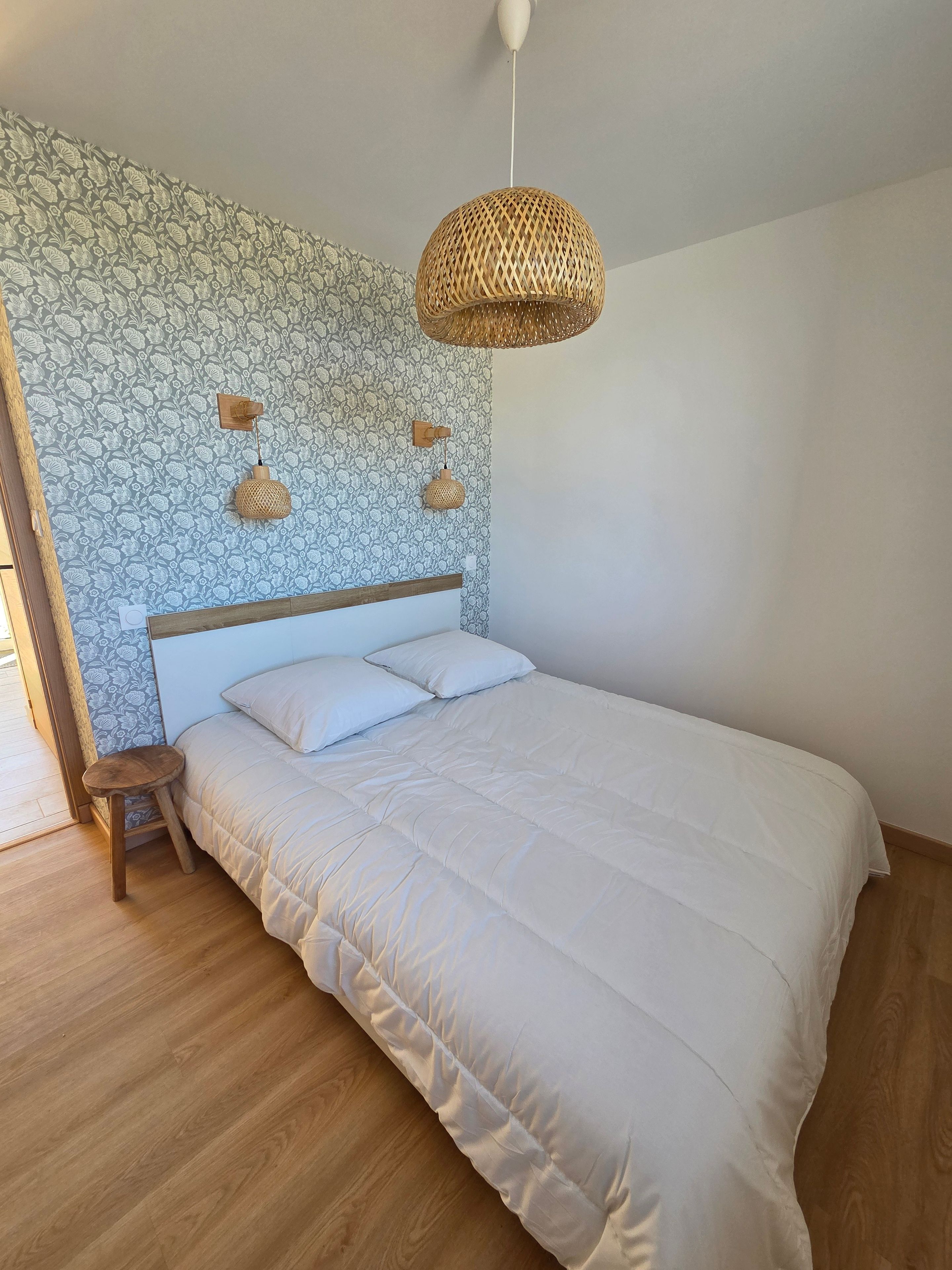 Een slaapkamer met een groot bed, een houten nachtkastje, een klein krukje, een structuurwand en een hanglamp.