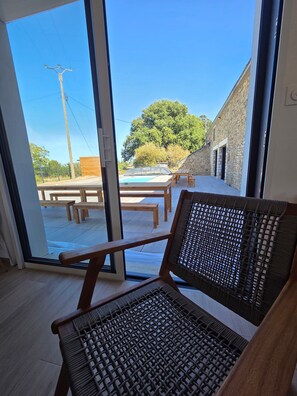 A room with a large sliding glass door leading to an outdoor area with a pool, wooden furniture, and a stone building. - Group Cottages Les Boizeul Pool - 2x15 Guests with Private Pool, Air Conditioning and Wi-Fi (Nivillac)
