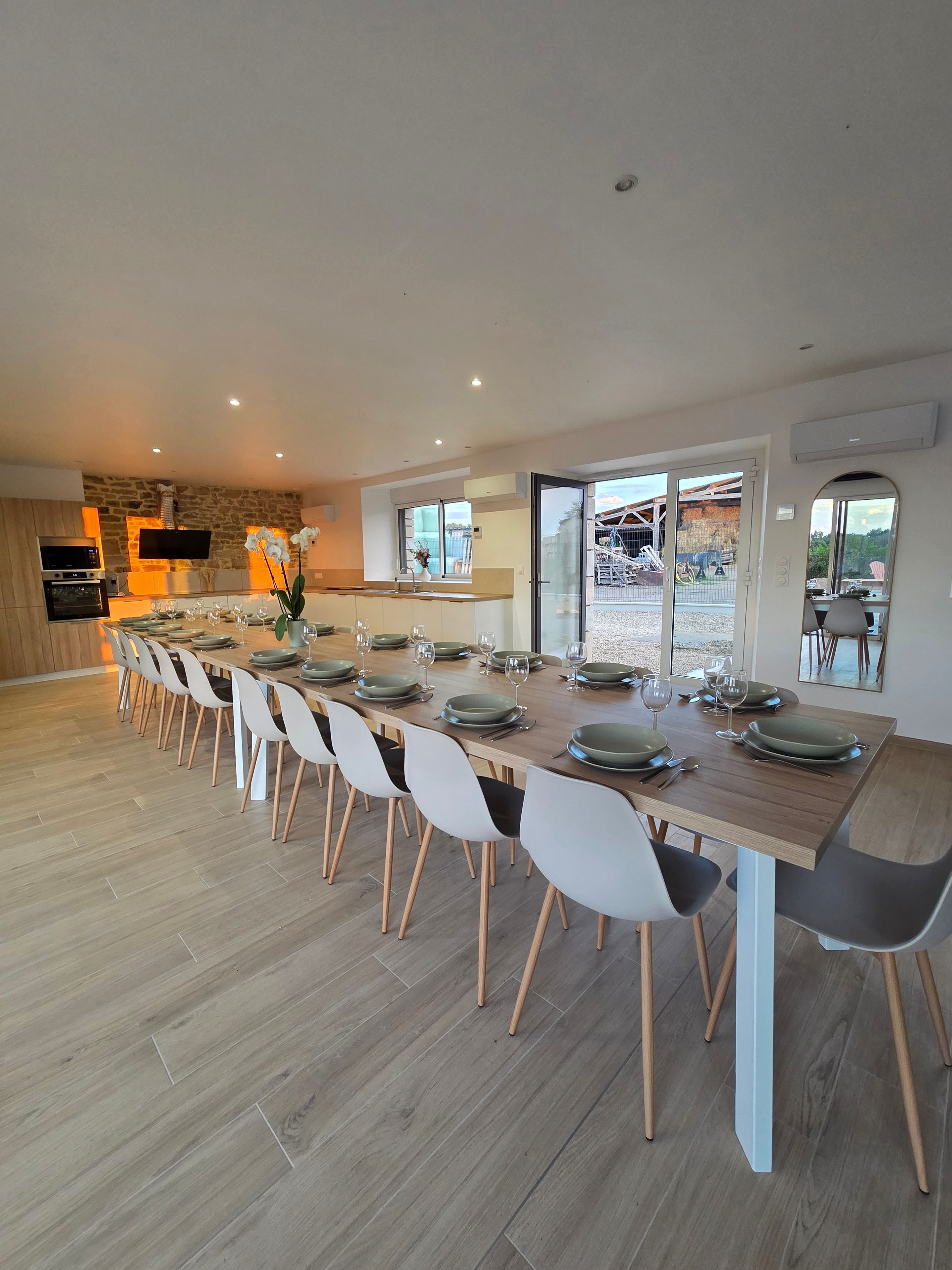 Un espace de restauration moderne avec une longue table dressée pour un repas, entourée de chaises, et une cuisine en arrière-plan.