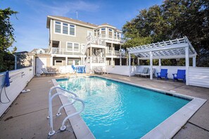 Une piscine avec une terrasse blanche, des chaises longues et un espace salon abrité.