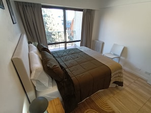 Een slaapkamer met een bed, een stoel, een raam met gordijnen en uitzicht op gebouwen.