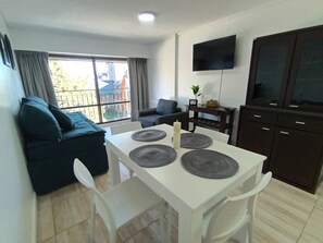 Una sala de estar moderna con un sofá, una mesa de comedor con individuales, un televisor de pantalla plana montado en la pared y vistas al exterior a través de amplios ventanales.