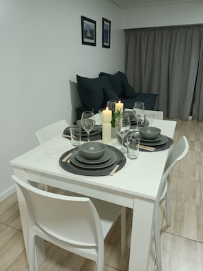 Een eettafel gedekt met borden, glazen en kaarsen, omringd door witte stoelen in een kamer met een grijze bank en gordijnen.
