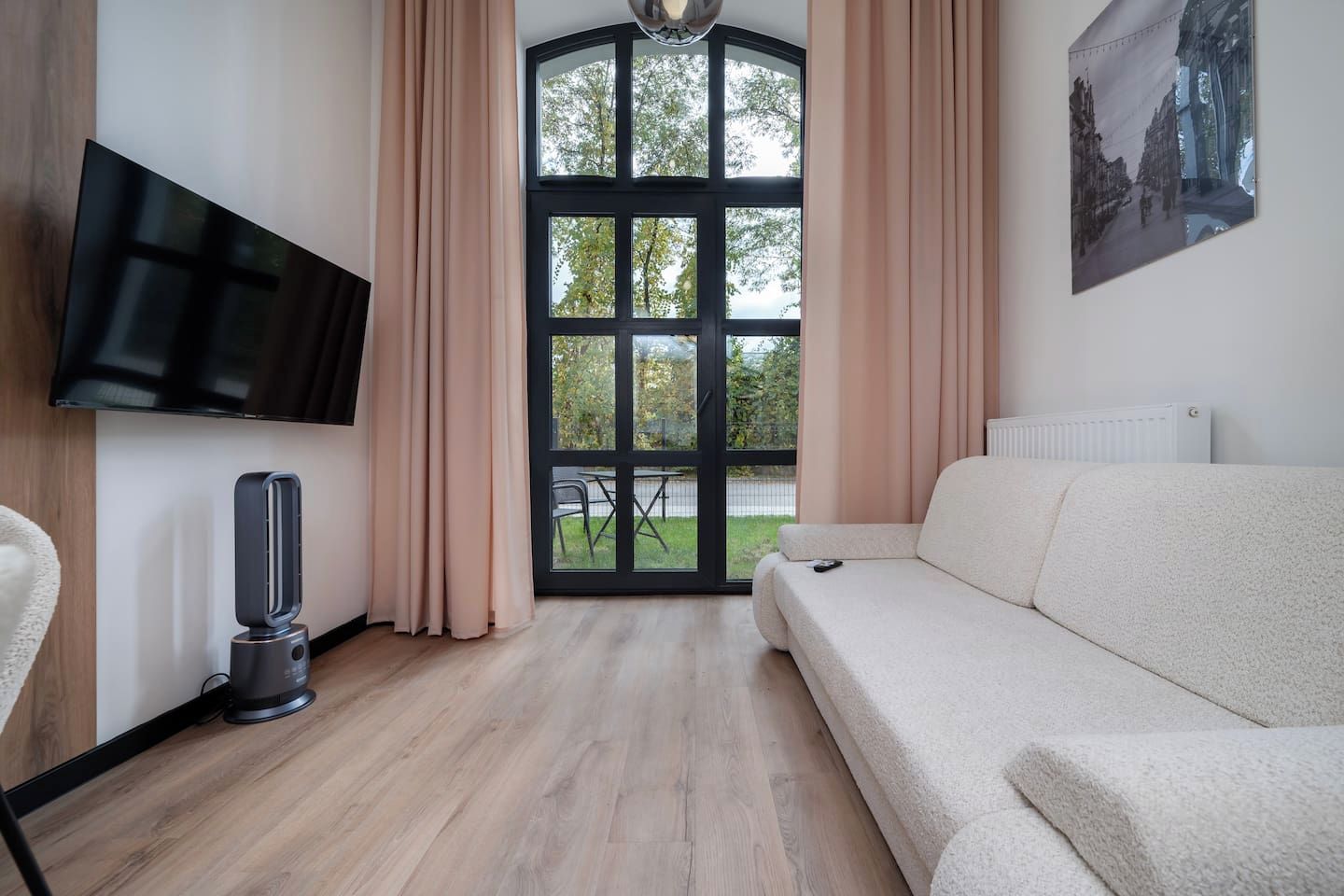 A modern living room with a sofa, a flat-screen TV, a vacuum cleaner, and a large window with curtains.