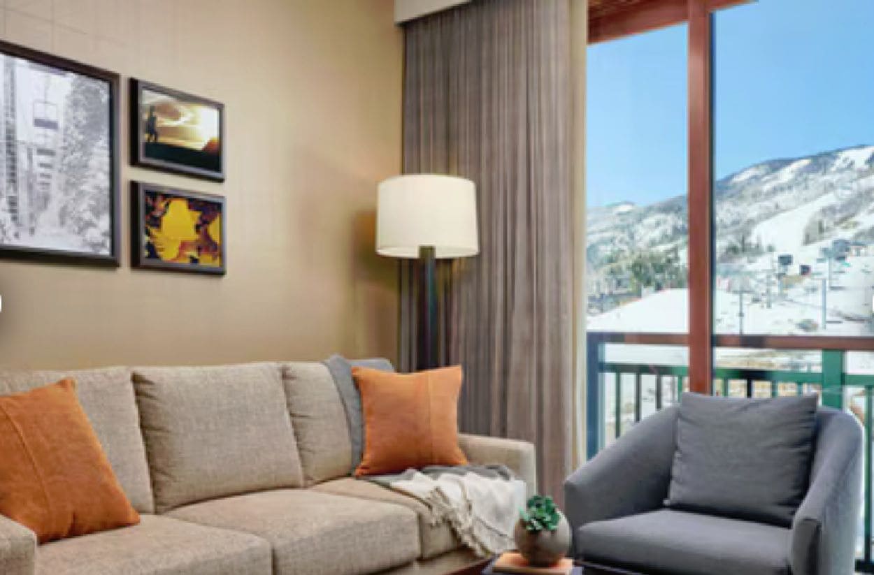 A living room with a sofa, armchair, and a lamp. There are framed pictures on the wall and a view of a snowy landscape through large windows.