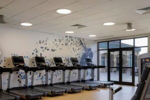 Un gimnasio con cintas de correr alineadas, un ventanal amplio y una pared con un diseño de mosaico.