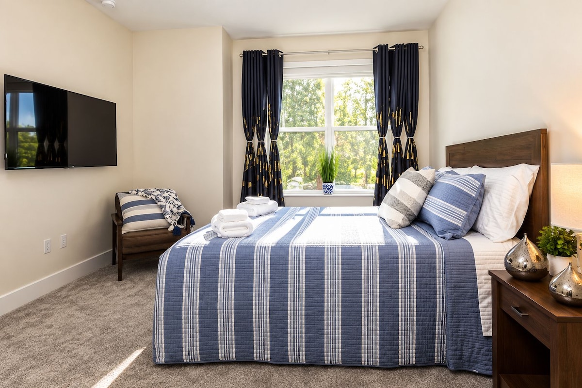 A bedroom with a bed, a chair, a TV, a window with curtains, and a bedside table with a lamp.