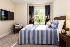 A bedroom with a bed, a chair, a TV, a window with curtains, and a bedside table with a lamp. - Clermont Sunshine Suites (Clermont)