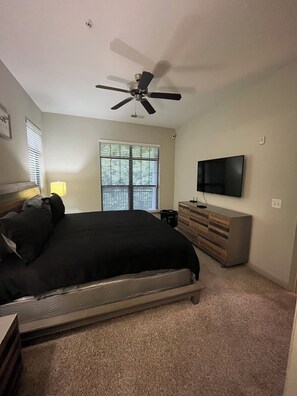 Une chambre à coucher comprenant un lit, un ventilateur de plafond, une fenêtre avec des stores, une commode et un téléviseur à écran plat.