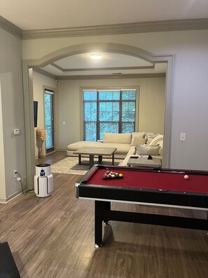 Une table de billard avec des billes, un canapé et une fenêtre avec des stores, dans une pièce dotée d’un plancher en bois et d’un plafond blanc.