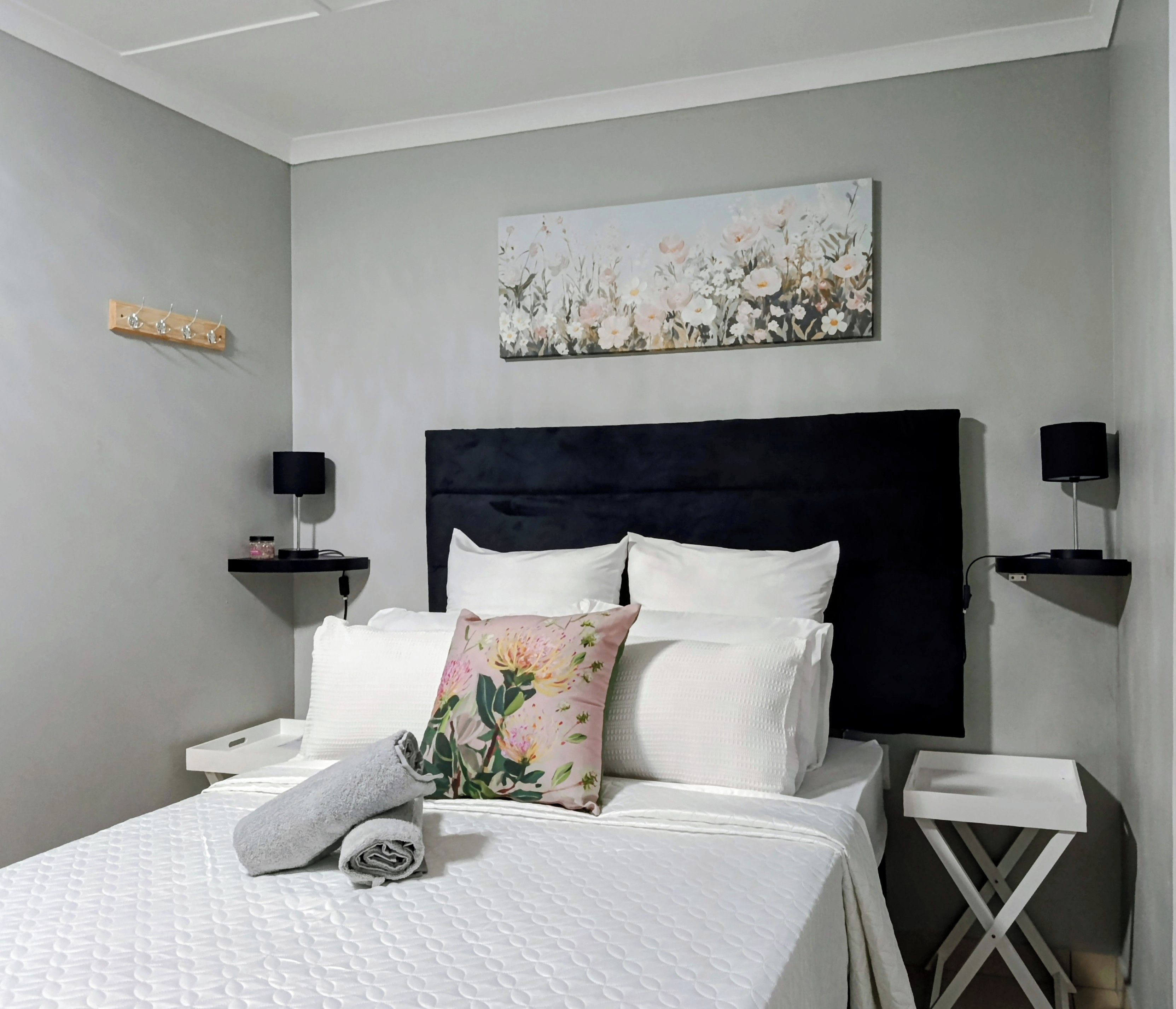 Un dormitorio con una cama grande, dos mesitas de noche, cabecera, una obra de arte en la pared y una pequeña estantería con lámpara.