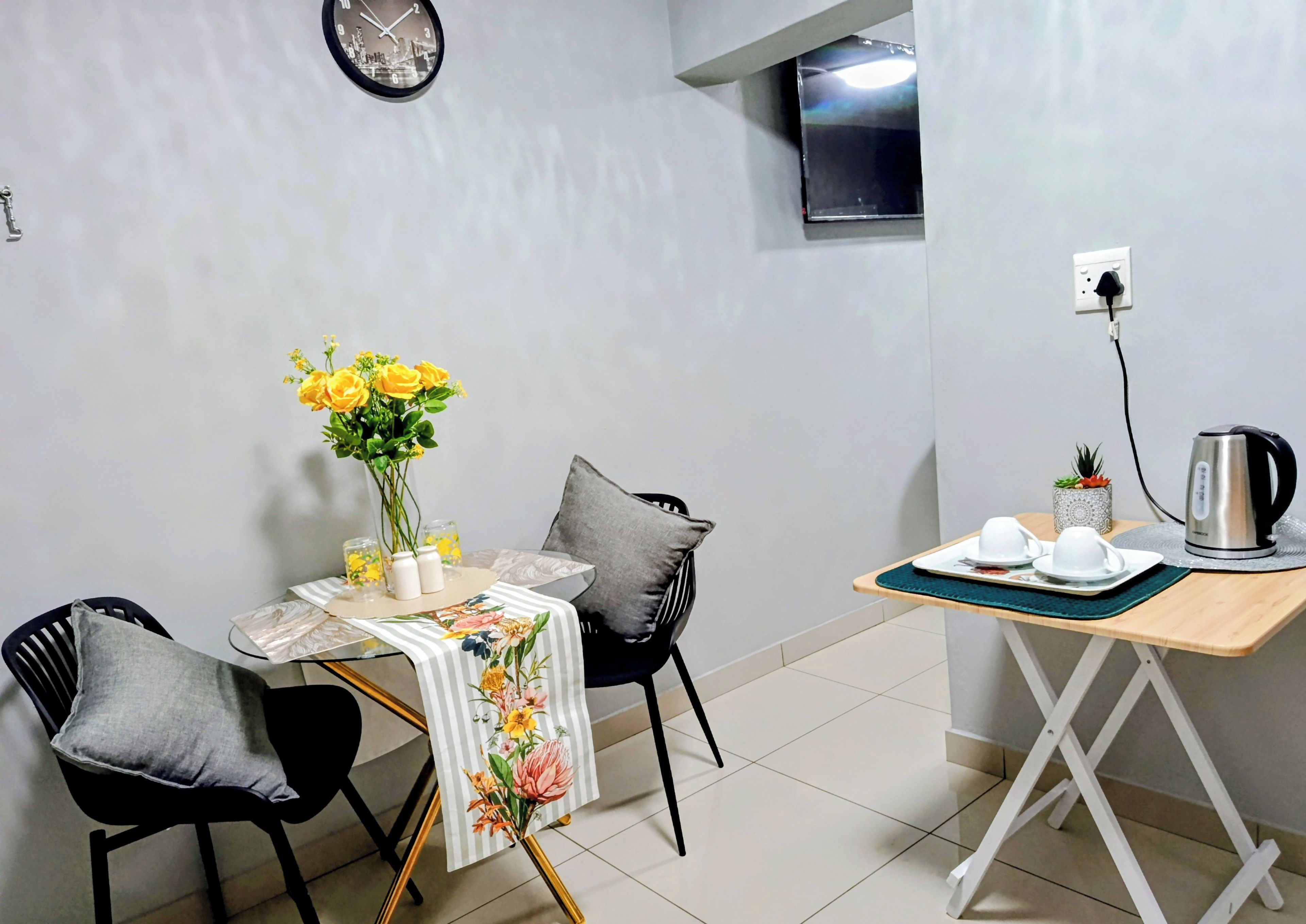 Un pequeño comedor con una mesa, sillas, un reloj, un televisor y un hervidor.