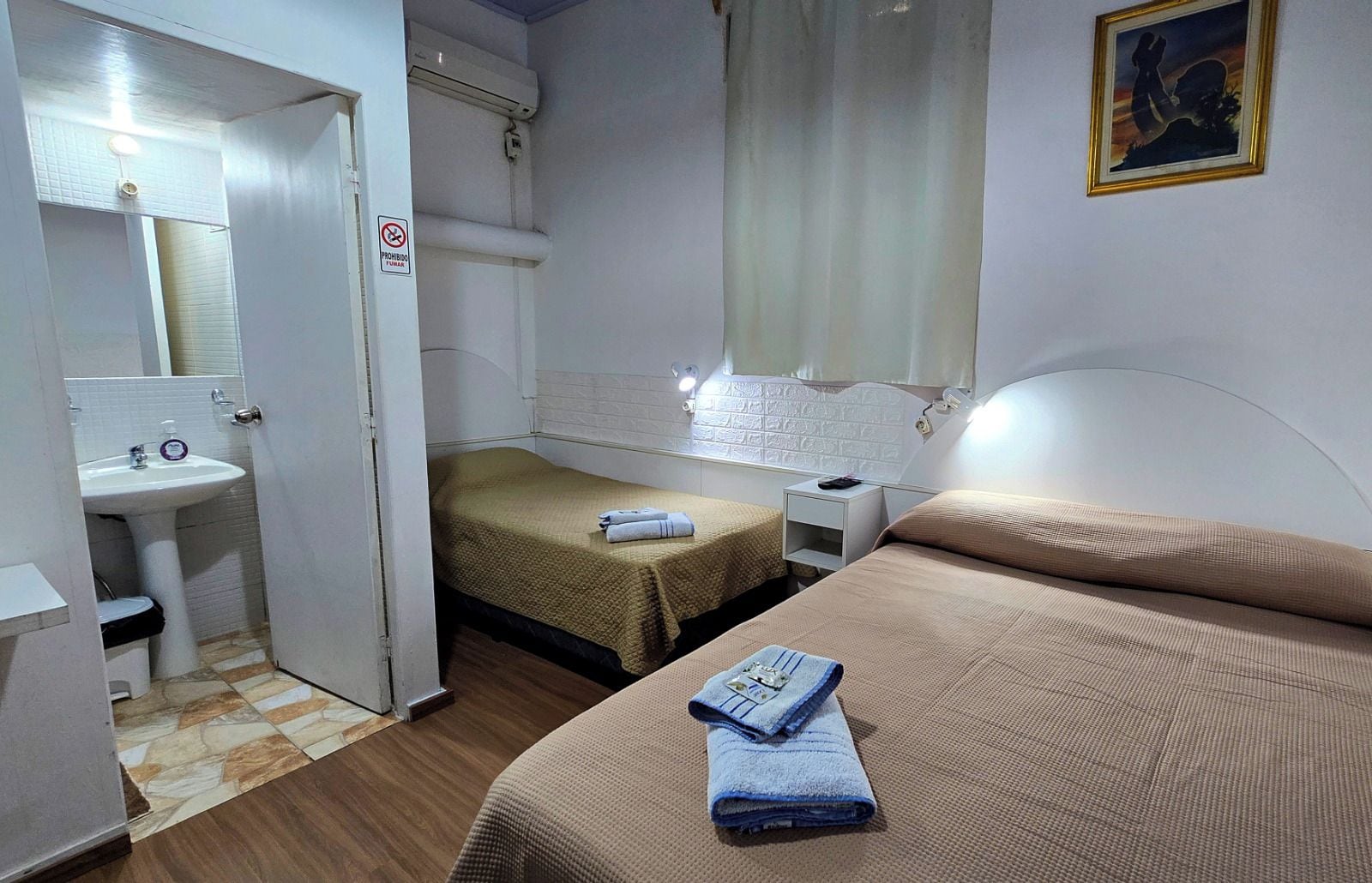 Una habitación con dos camas, una mesita de noche, un tocador, un ventanal y un baño con lavabo y espejo.