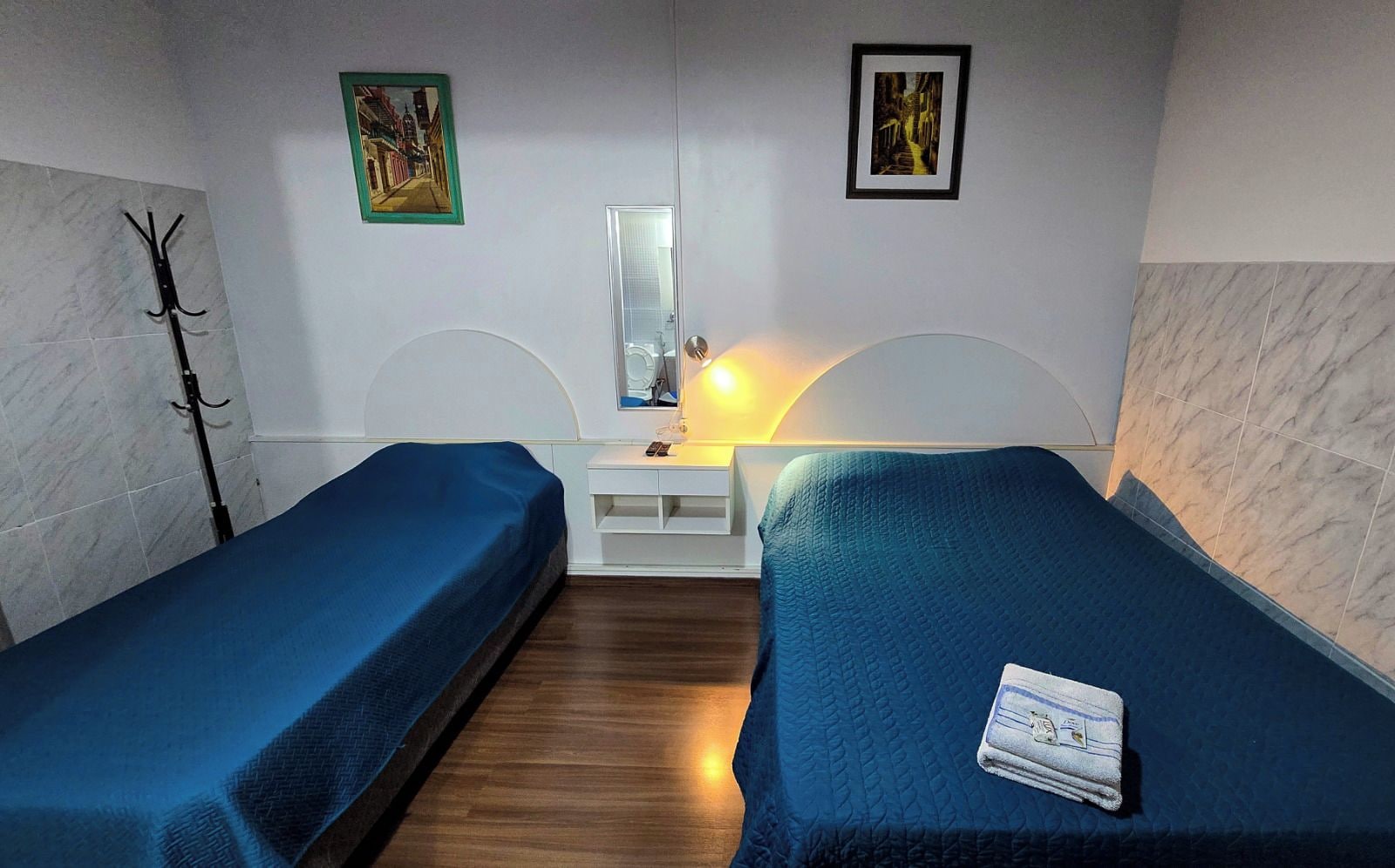 Una habitación con dos camas individuales, una mesita de noche con lámpara, un perchero y dos cuadros en la pared.