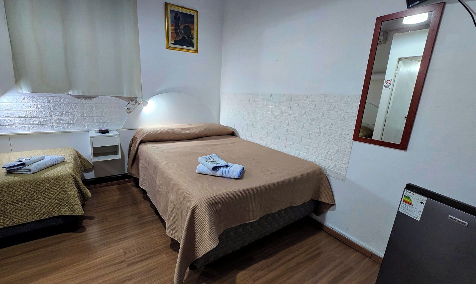 Habitación de hotel con dos camas, una mesita de noche, una lámpara, un espejo y un refrigerador.