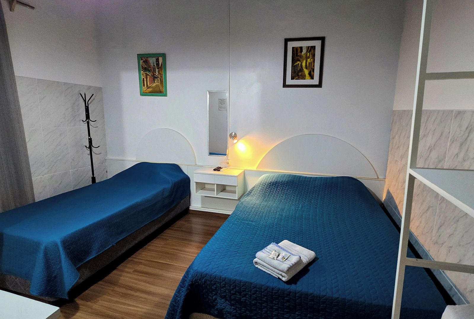 A room with two beds, a bedside table with a lamp, a coat rack, and two framed pictures on the wall.