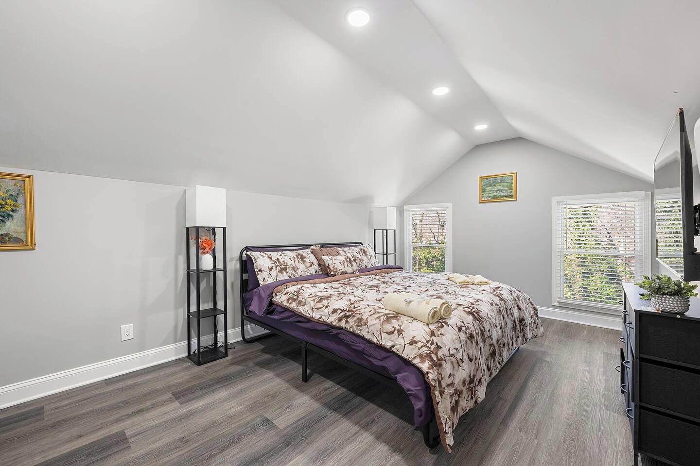 Une chambre à coucher comprenant un lit, une commode, une table de chevet, une fenêtre avec des stores et un plafond doté d’un éclairage encastré.