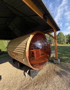 A cylindrical wooden structure with a transparent side, supported by a metal roof and wooden beams, situated outdoors. - Historic Wiseman Retreat (Newland)