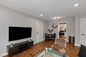 A modern living room with a flat-screen TV, dark wood flooring, a dining area with a table and chairs, and a kitchen in the background. - Prime Location Midtown Omaha (Omaha)