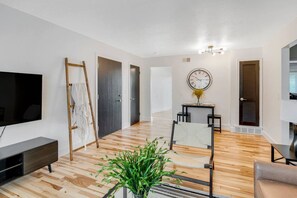 Una sala de estar moderna con un televisor de pantalla plana, piso de madera, un cuadro en la pared, un reloj y una planta.