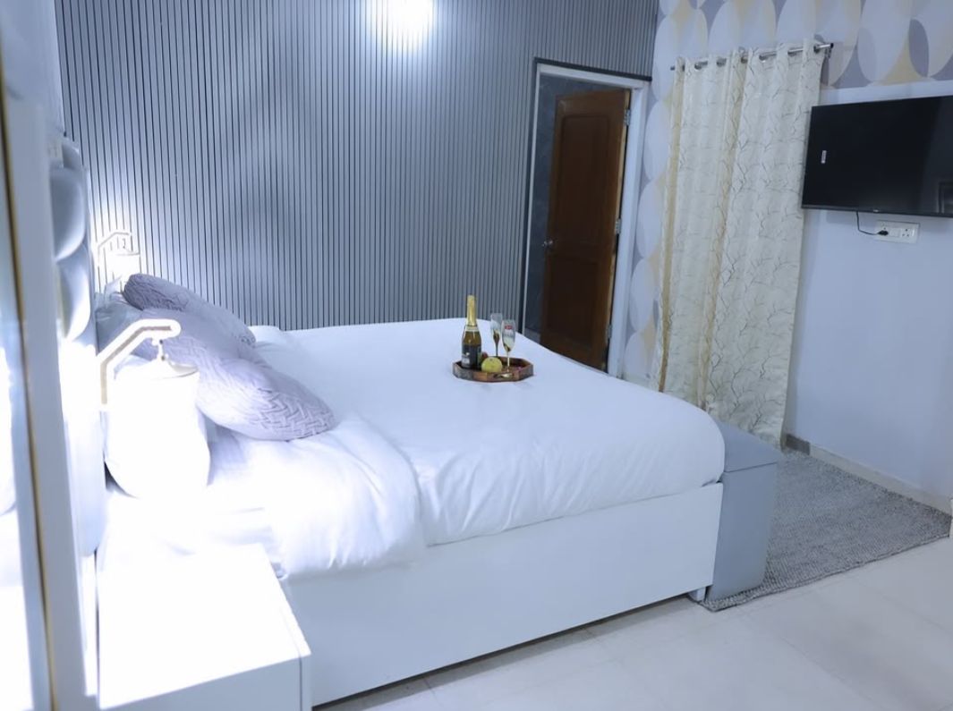 Une chambre d’hôtel avec un lit, une table de chevet sur laquelle se trouvent une bouteille et des verres, un téléviseur fixé au mur et une porte.