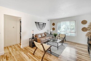A spacious living room with a sofa, coffee table, and chairs, a large window with blinds, and a wooden floor. - Prime Location! HGTV Design! (Omaha)