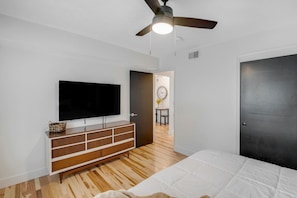 Quarto com cama, televisão fixada na parede, cômoda de madeira, ventilador de teto e porta que dá para outro ambiente.