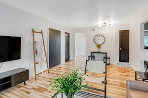 A modern living room with a flat-screen TV, a wooden floor, a dining area with a table and chairs, and a large wall clock. - Prime Location! HGTV Design! (Omaha)