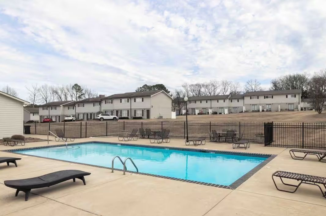 A swimming pool with lounge chairs, a fenced area, and a row of buildings in the background.
