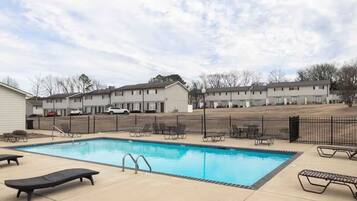 A swimming pool with lounge chairs, a fenced area, and a row of buildings in the background.