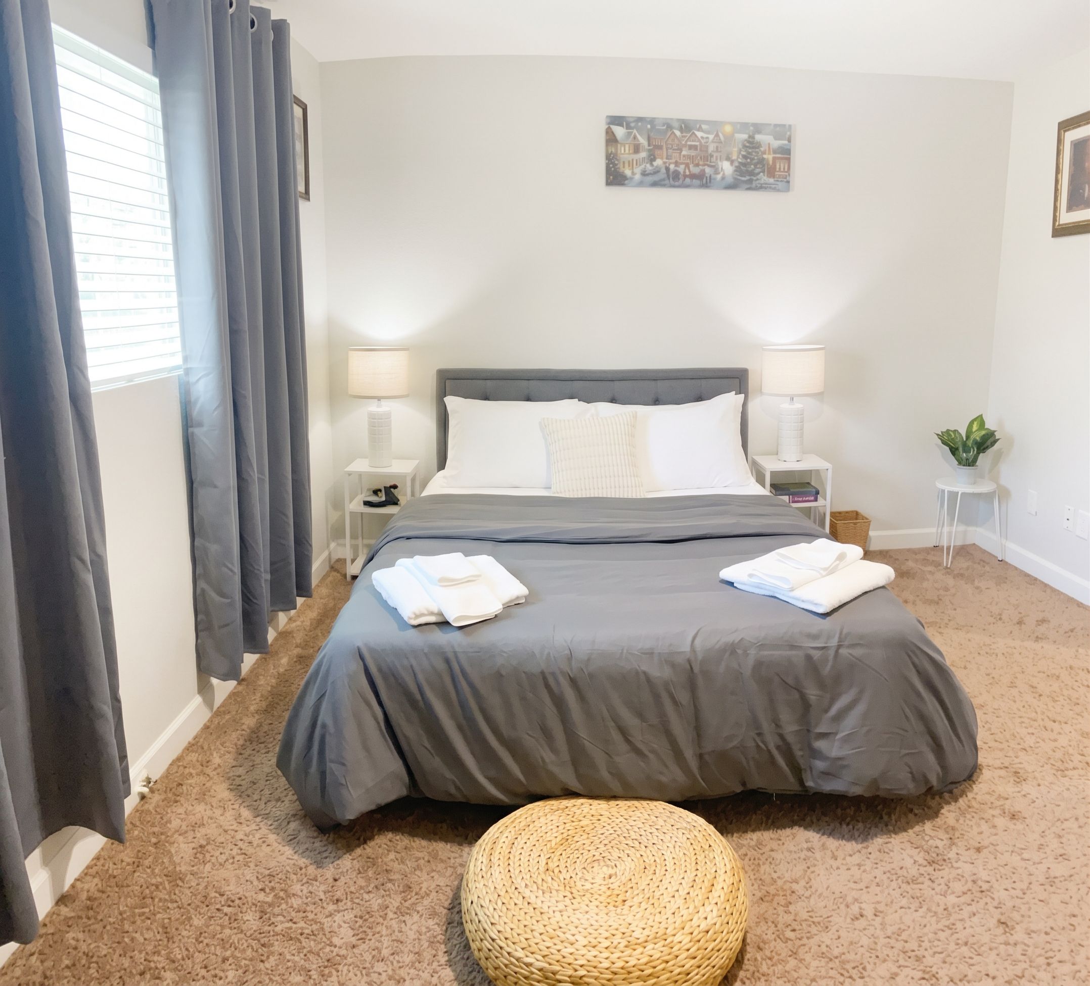 A bedroom with a bed, two bedside tables, a lamp on each, a painting on the wall, and a small plant on a side table.