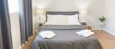 A bedroom with a bed, two bedside tables, a lamp on each, a painting on the wall, and a small plant on a side table.