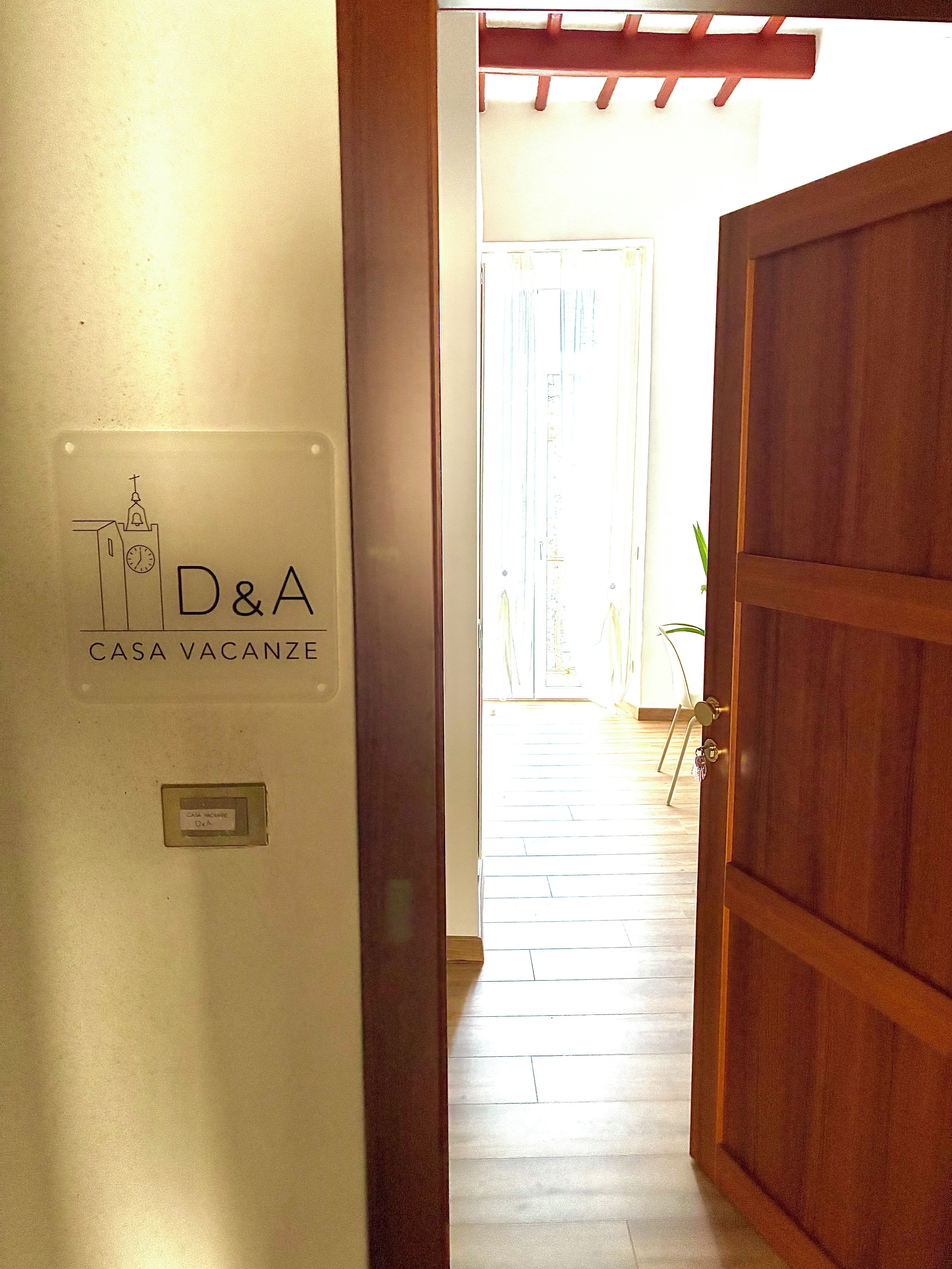 Un pasillo que conduce a una habitación con puerta de madera, un cartel que indica 'D&A CASA VACANZE', y vista de una habitación luminosa con una planta.