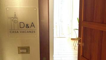 Un pasillo que conduce a una habitación con puerta de madera, un cartel que indica 'D&A CASA VACANZE', y vista de una habitación luminosa con una planta.