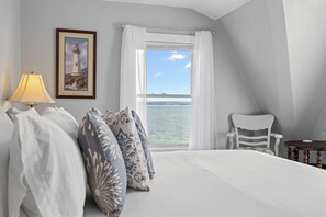 Une chambre à coucher avec un lit, des oreillers, une lampe, une chaise, une fenêtre avec des rideaux et un tableau encadré au mur.