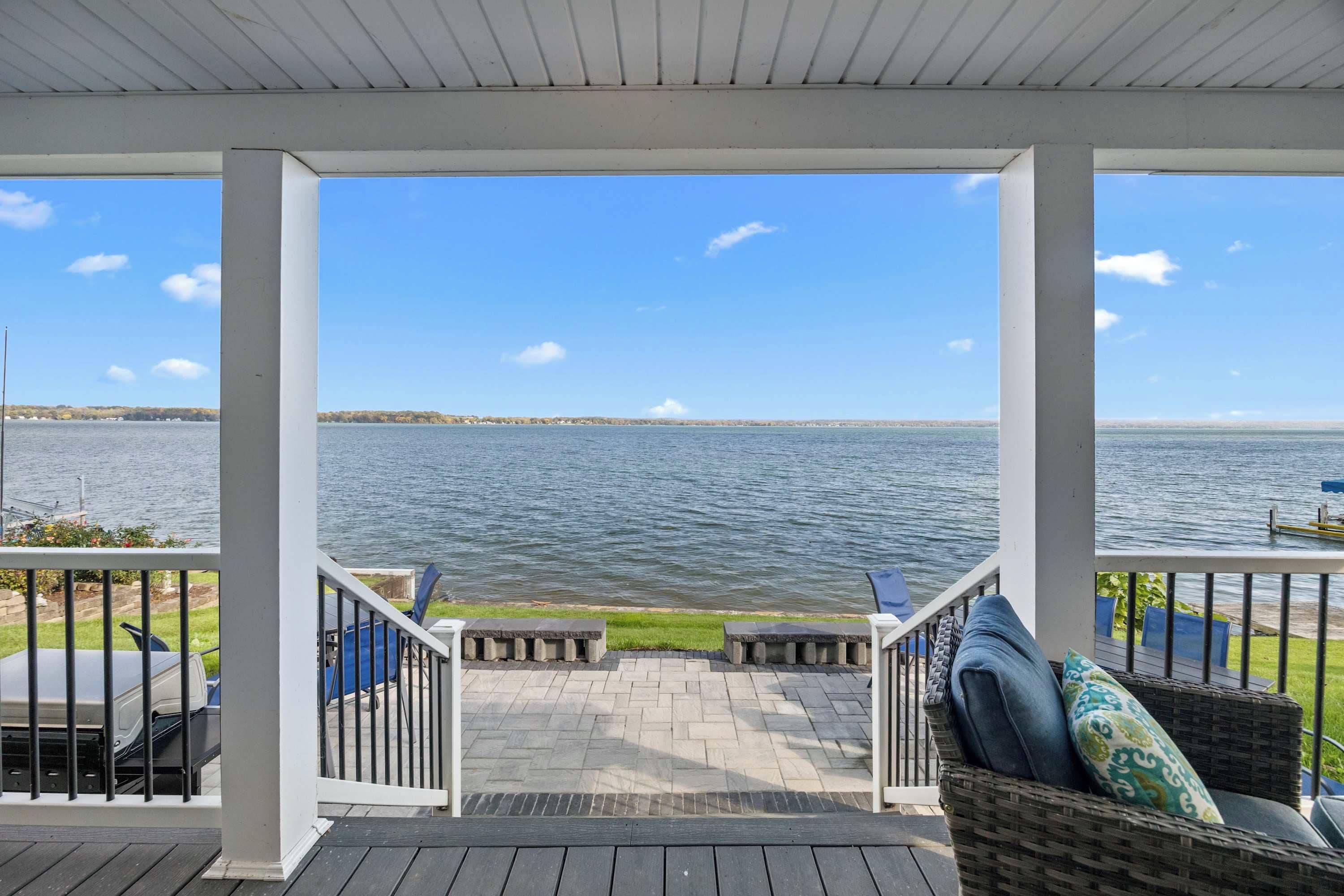A covered outdoor area with a view of a lake, featuring a wooden deck, outdoor furniture, and a railing.